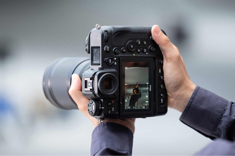 Photographer holding a professional cinema camera with attached lens, viewed from behind against an overcast sky.