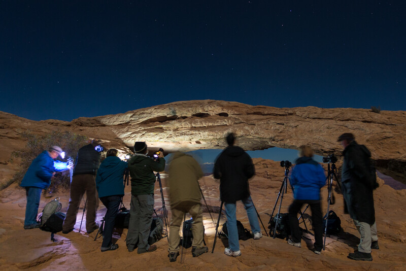Group of photographers with tripods and cameras positioned on rocky terrain facing an illuminated cliff face at night.