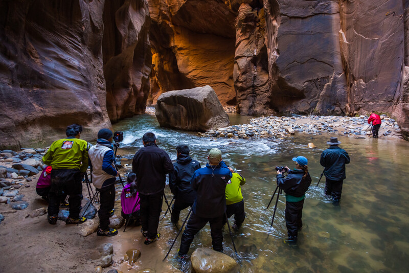Group of hikers wading through a shallow river in a narrow canyon with towering rock walls.