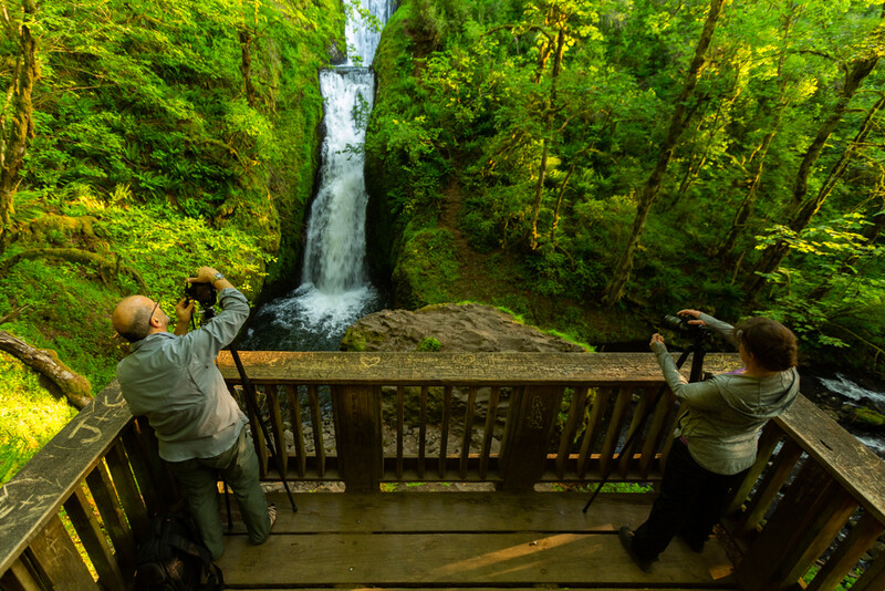 Two photographers on a wooden deck overlooking a waterfall framed by dense green forest.