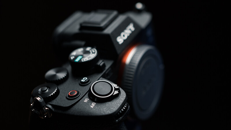 Close-up of a professional DSLR camera body with control dials and buttons in sharp focus against a blurred black background.