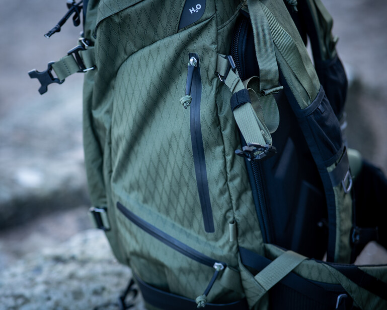 Close-up of a sage green hiking backpack with multiple compartments and straps against a blurred outdoor background.