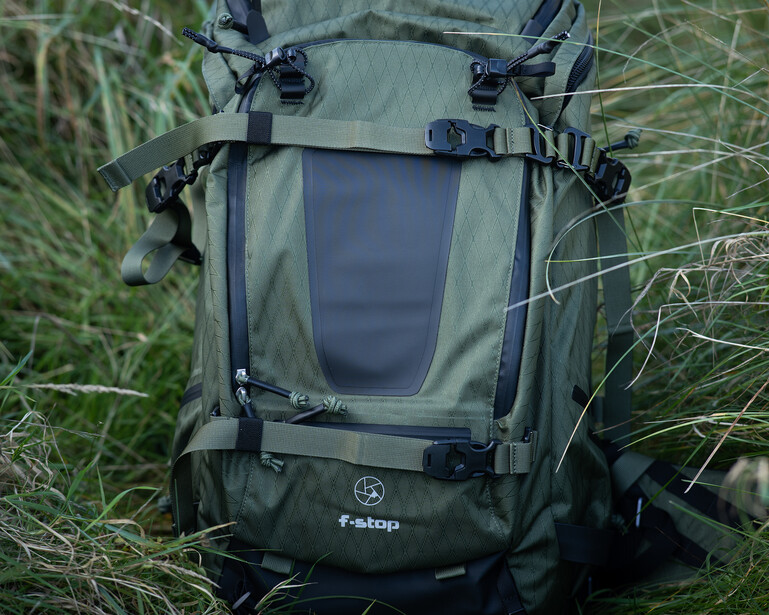 Olive green waterproof backpack with dark panel displayed on grass, showing front view with carry handles and attachment points.