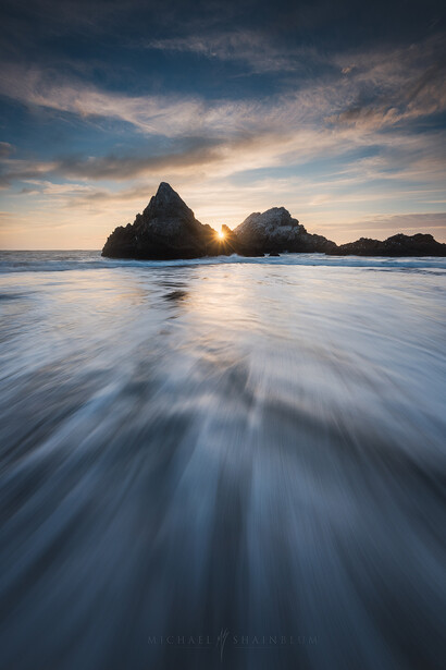 Rocky sea stacks silhouetted against a golden sunset over ocean waves with motion blur.