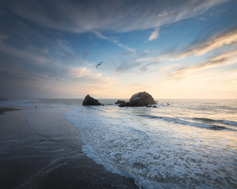 Rocky sea stacks rising from the ocean during golden hour with waves rolling onto a dark sand beach.