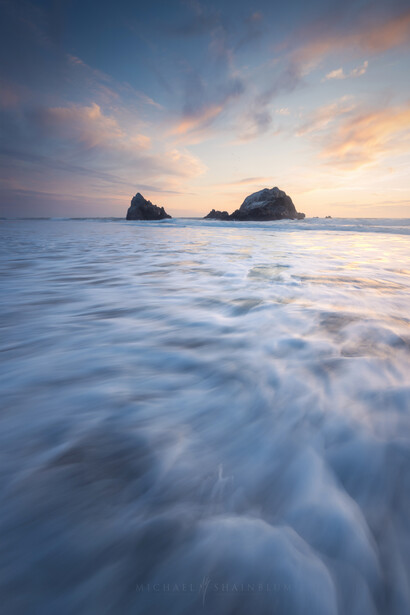Rocky sea stacks silhouetted against a golden sunset over a long-exposure beach.