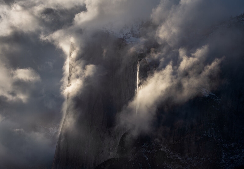 Dramatic waterfall cascading through layered clouds and mist in mountainous terrain.