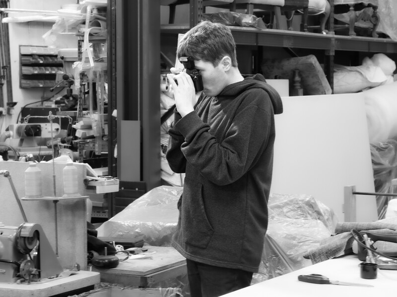 Photographer holding camera to face while working in studio with shelving and equipment visible in background.