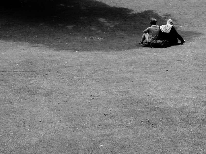 Two figures sitting close together on an empty asphalt surface with dramatic shadow.