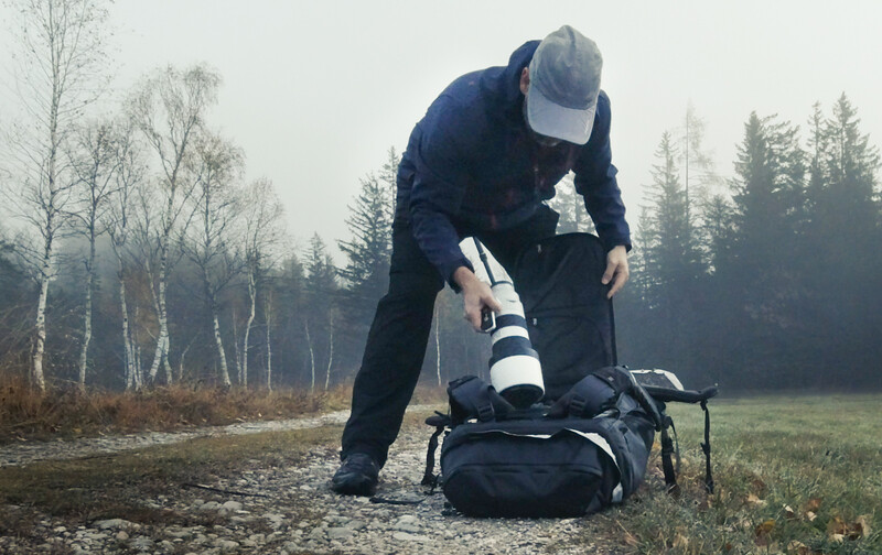 Photographer bending over a camera bag in an outdoor forest setting, adjusting equipment.