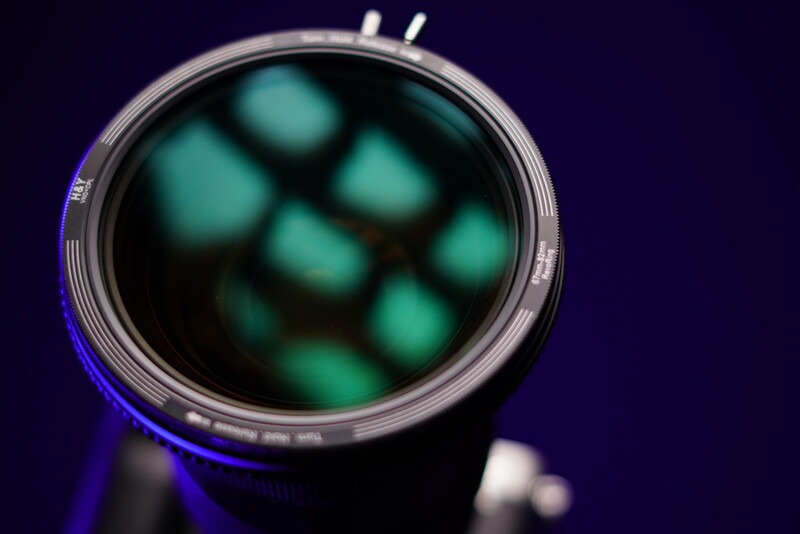 Close-up of a camera lens front element showing green-tinted dust spots and internal reflections against a dark background.