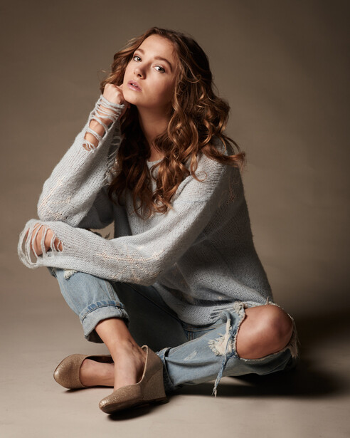 Woman in gray sweater and distressed jeans sitting cross-legged against neutral background.