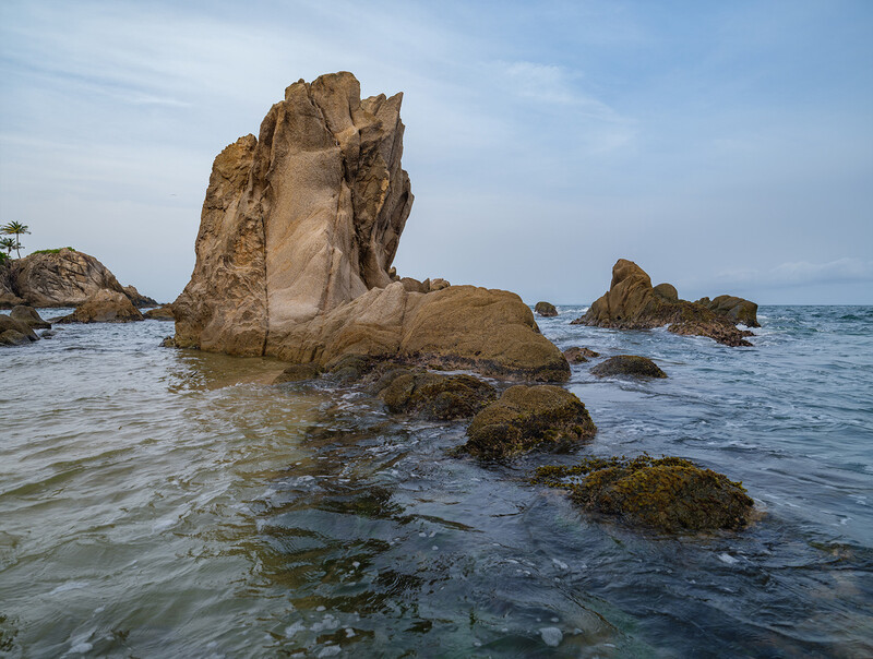 Tall granite rock formation rising from shallow coastal water surrounded by moss-covered boulders.