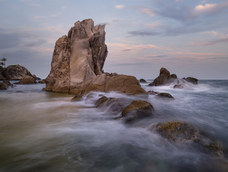 Tall granite rock formation rising from turbulent ocean waves at dusk, surrounded by smaller boulders.