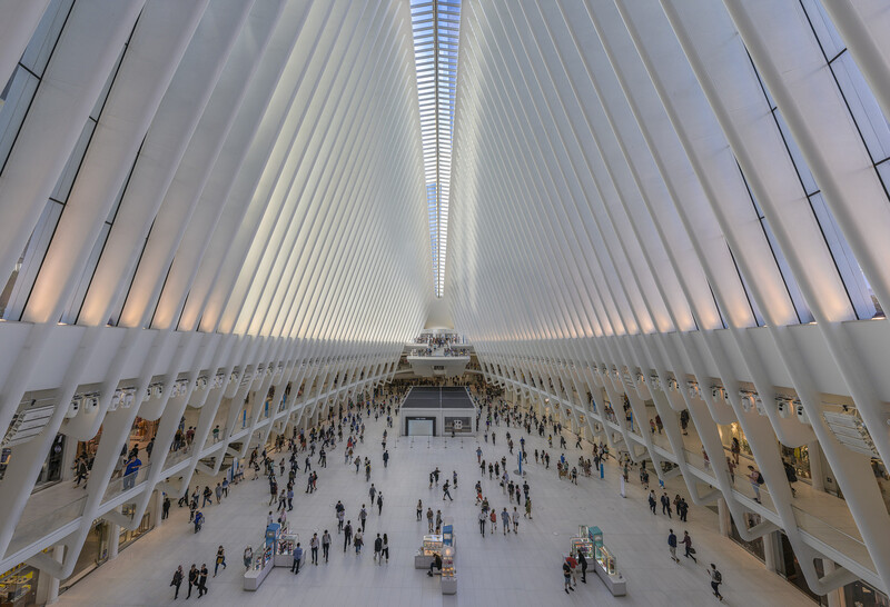 Interior of a modern white architectural space with symmetrical ribbed ceiling, multiple levels, and crowds of people on the main floor.