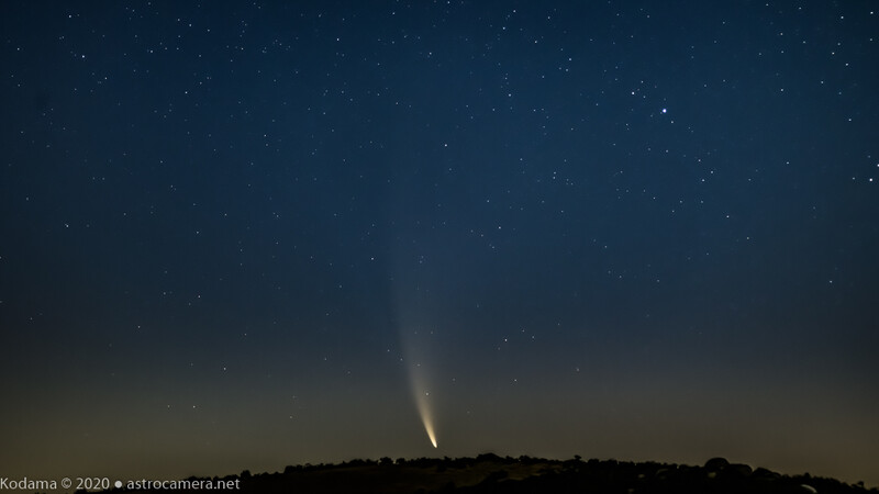 Bright comet with distinct tail rising above dark silhouetted landscape against starry night sky.