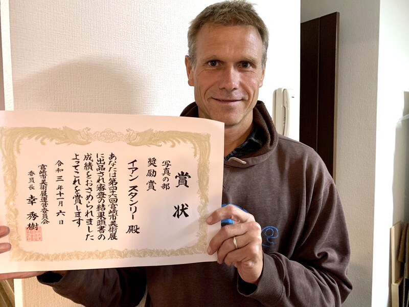 Man holding a Japanese calligraphy certificate with text and red stamp seal.
