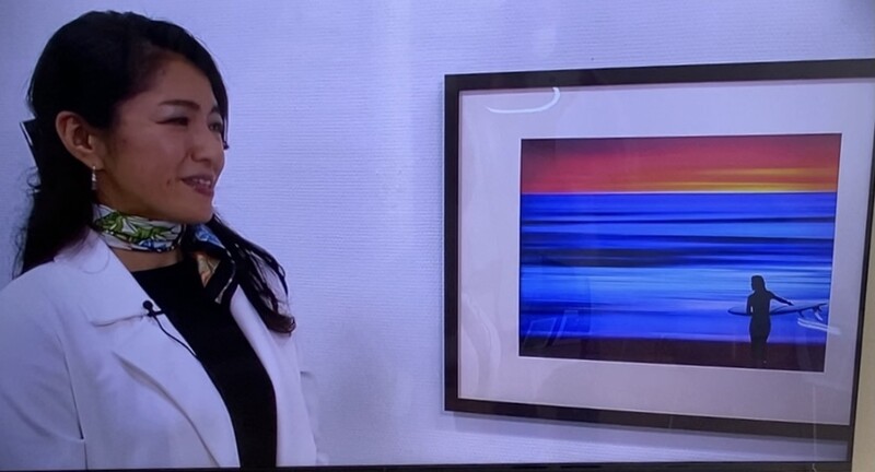 Woman in white blazer viewing framed photograph of sunset seascape with silhouetted figure.