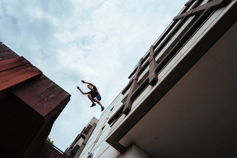Parkour athlete mid-leap between two modern buildings against cloudy sky.