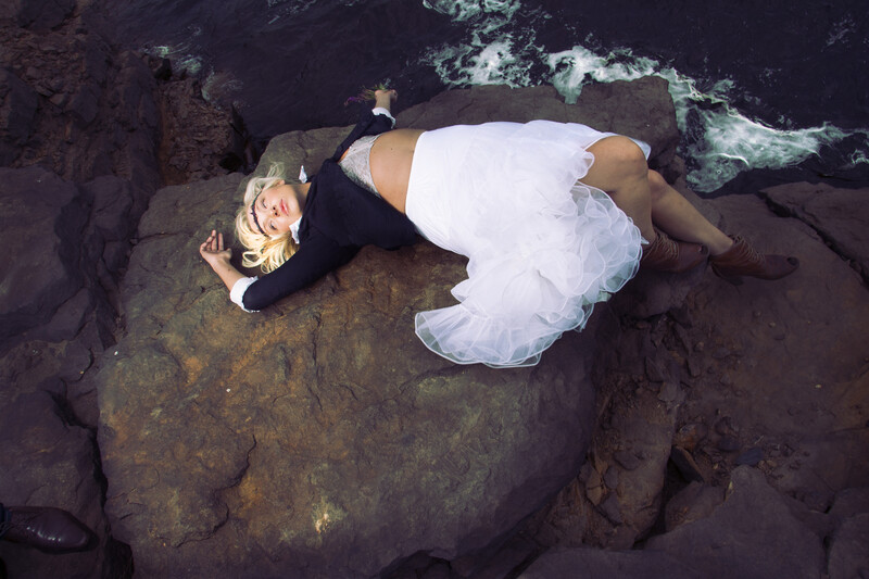 Woman in white dress lying on dark volcanic rock formations near ocean waves.