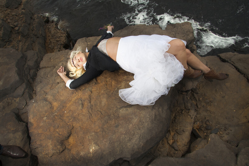 Woman in white dress lying on dark rocky coastal formations with ocean waves visible in background.