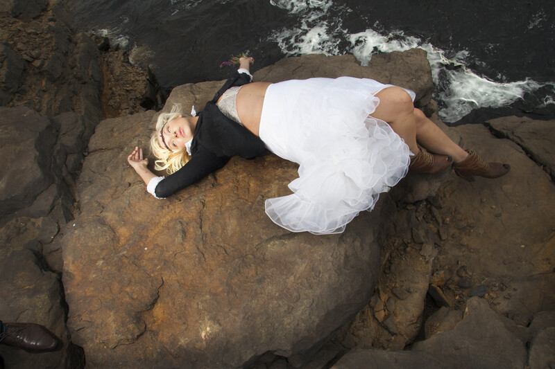 Woman in white dress lying on dark rocky coastal formation with ocean waves in background.