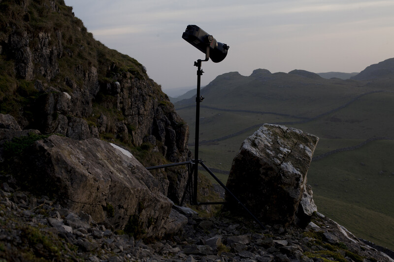 Professional lighting rig mounted on tripod positioned on rocky mountain outcrop overlooking valley.