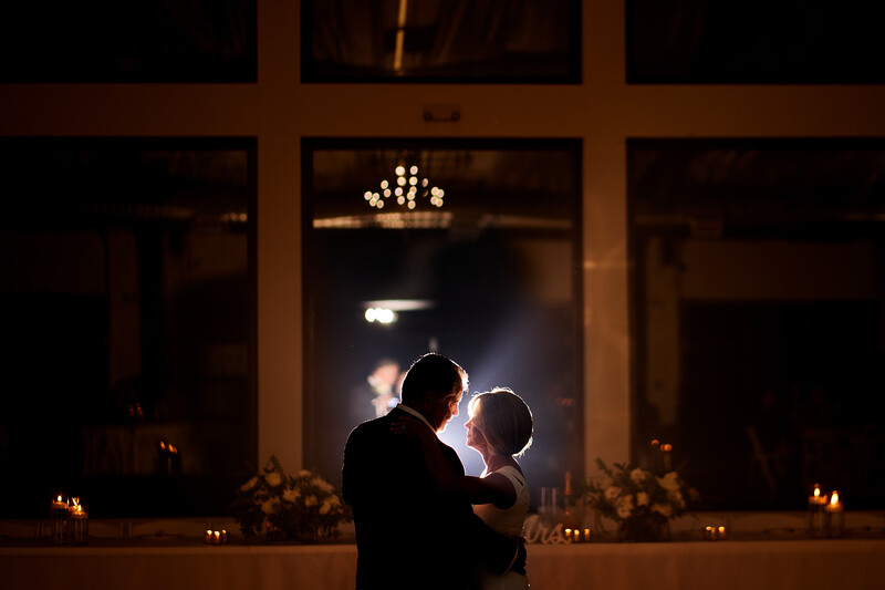 Silhouetted couple dancing in front of large windows with chandelier and candles visible in background.