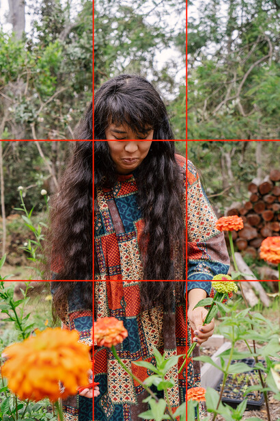 Person with long dark hair wearing patterned clothing examining orange flowers in a garden setting.