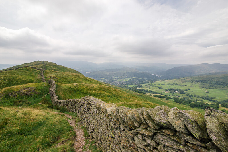 Stone wall running along a grassy hillside with a valley and distant mountains visible under overcast skies.