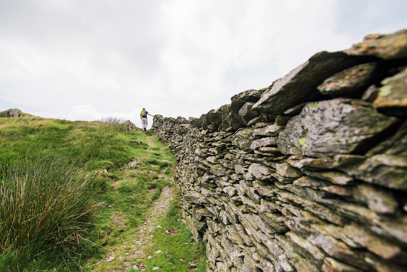 Stone wall in sharp focus alongside a grassy hiking trail with a distant hiker on the hillside.