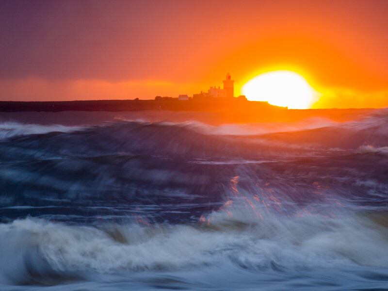 Waves crash on a beach at sunset with a bright sun low on the horizon and a coastal city silhouetted against the orange sky.