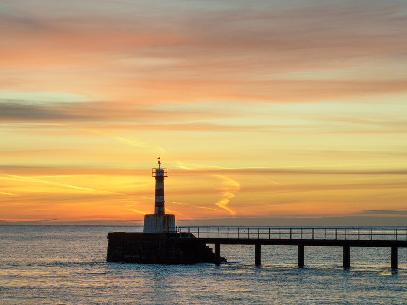 Lighthouse silhouetted against golden sunset sky with connecting pier extending into calm water.
