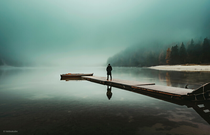 Solitary figure standing on a fallen log extending into a misty lake surrounded by evergreen forest.