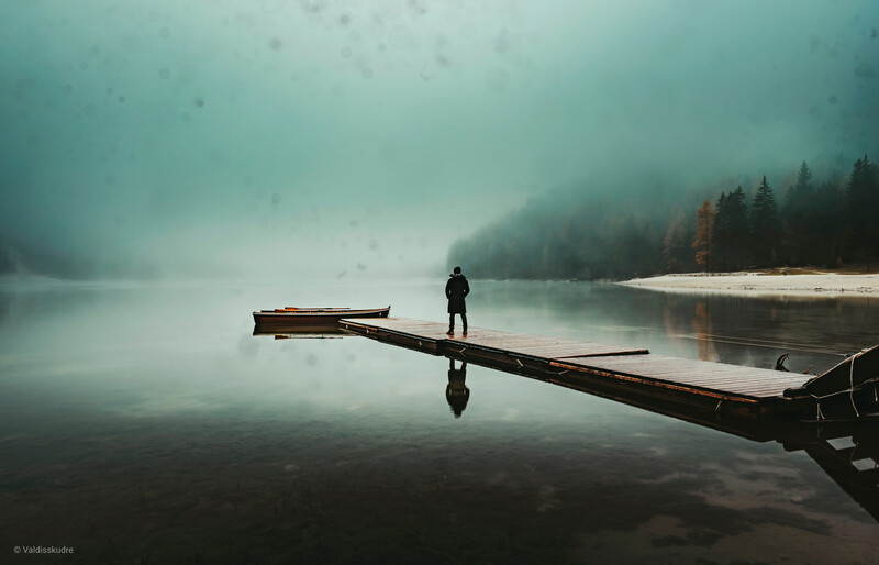 Solitary figure standing on a fallen log extending into a misty lake surrounded by forested shores.