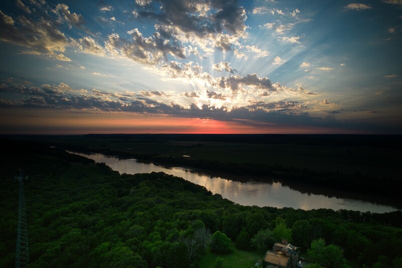 River winding through dense green forest at sunset beneath dramatic cloudy sky with golden light breaking through.