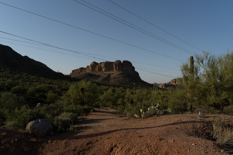 Desert landscape with rocky butte formation, saguaro cacti, and dirt road beneath clear sky.