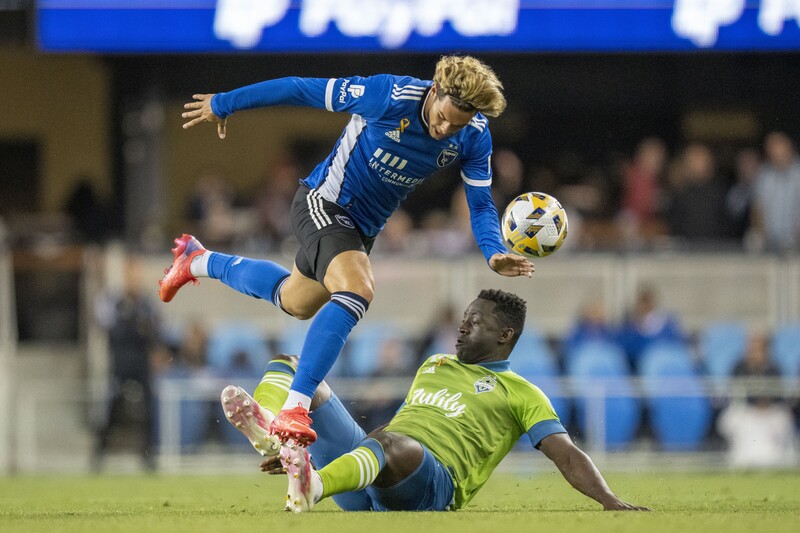 Soccer player in blue jersey leaps over sliding defender in yellow-green kit during match.