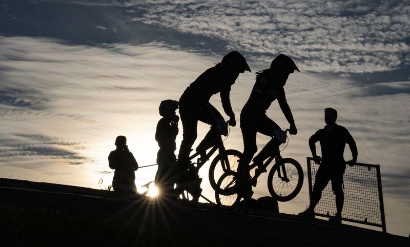 Silhouetted figures with bicycles and a soccer goal against a bright sky at sunset or sunrise.