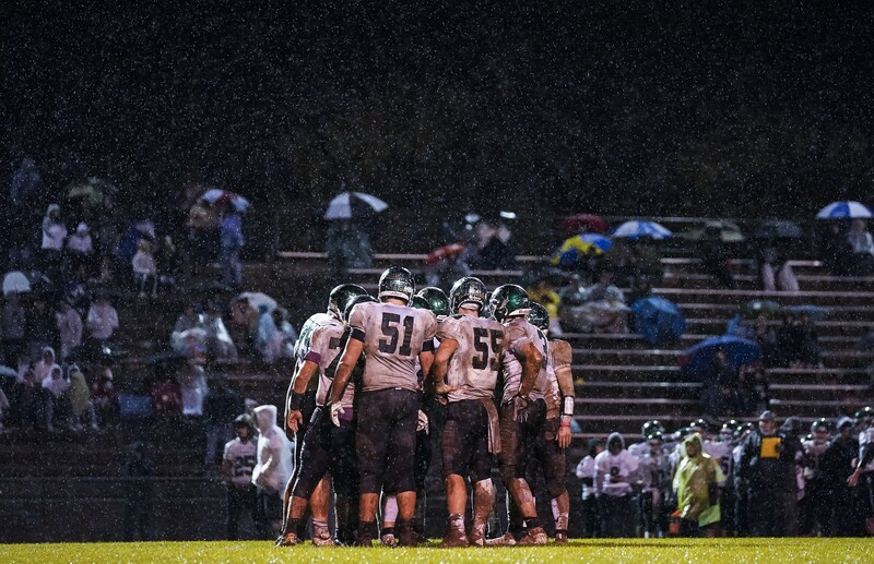 Football team huddled together in rain during night game with spectators in bleachers behind them.