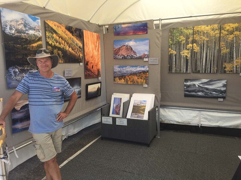 Photographer standing in front of displayed landscape photographs at an outdoor exhibition tent.