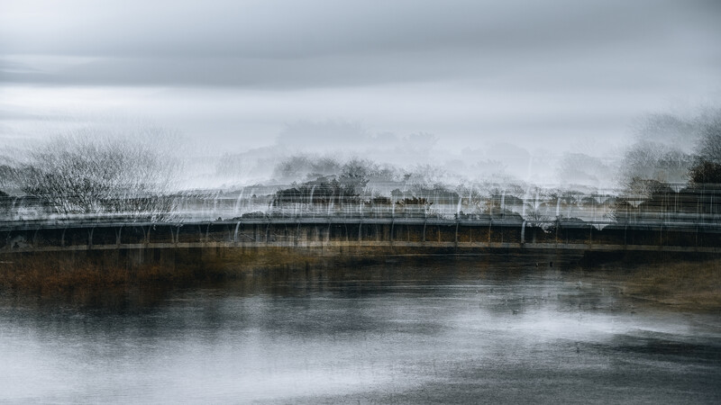 Industrial waterfront with bridge and bare trees reflected in still water under overcast sky.