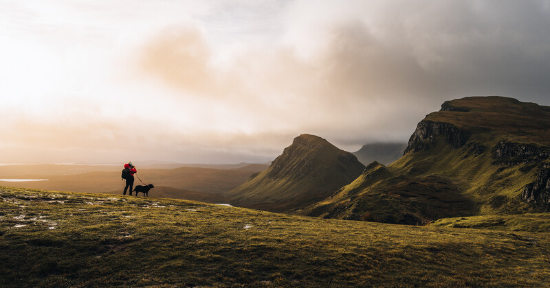 Hiker and dog on moss-covered terrain with dramatic basalt mountains in the distance under overcast sky.