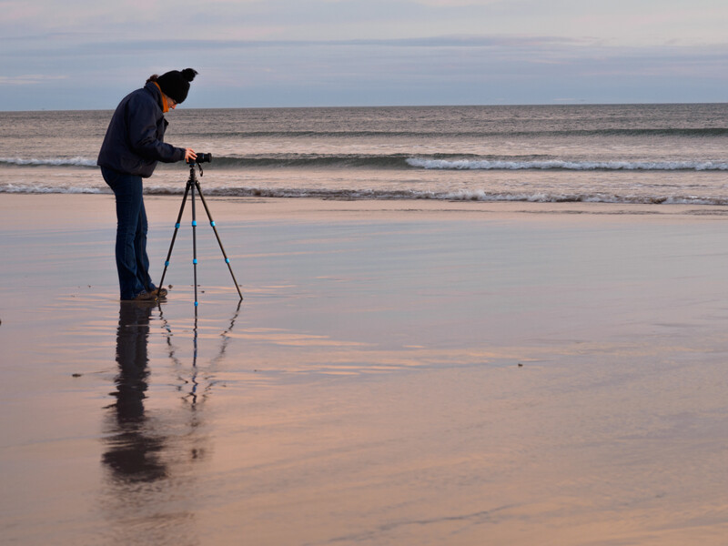 Photographer on a wet beach at low tide, bent over a tripod-mounted camera facing the ocean at twilight.