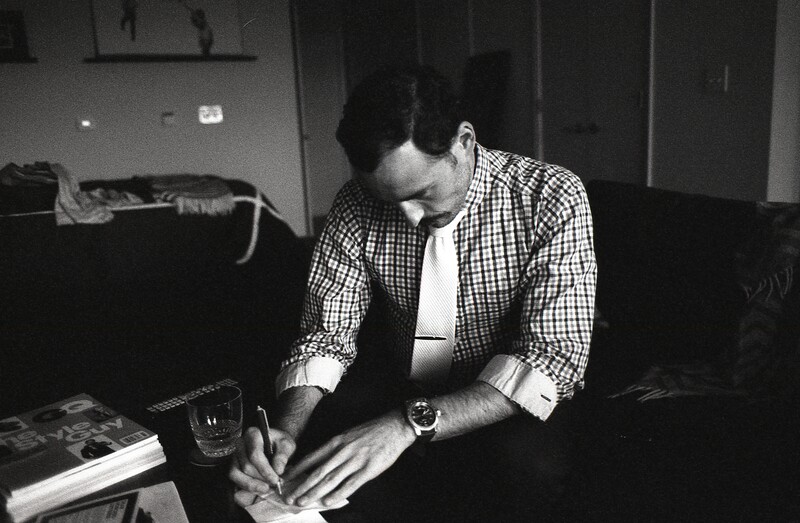 Man in checkered shirt writing or sketching at a desk in a black and white photograph.