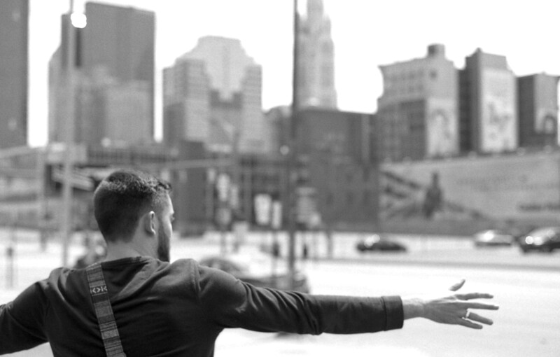 Man with outstretched arm facing urban skyline in black and white photograph.