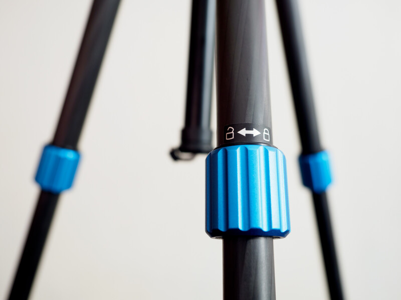 Blue tripod spreader with width measurement indicator mounted on black tripod legs against neutral background.