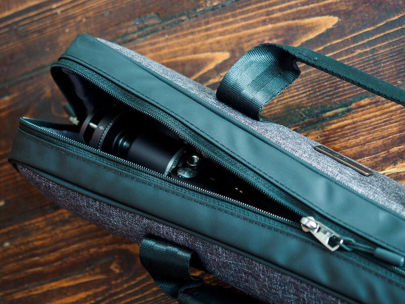 Black camera lens and lens cap partially emerging from an open gray padded gear bag on a wooden surface.