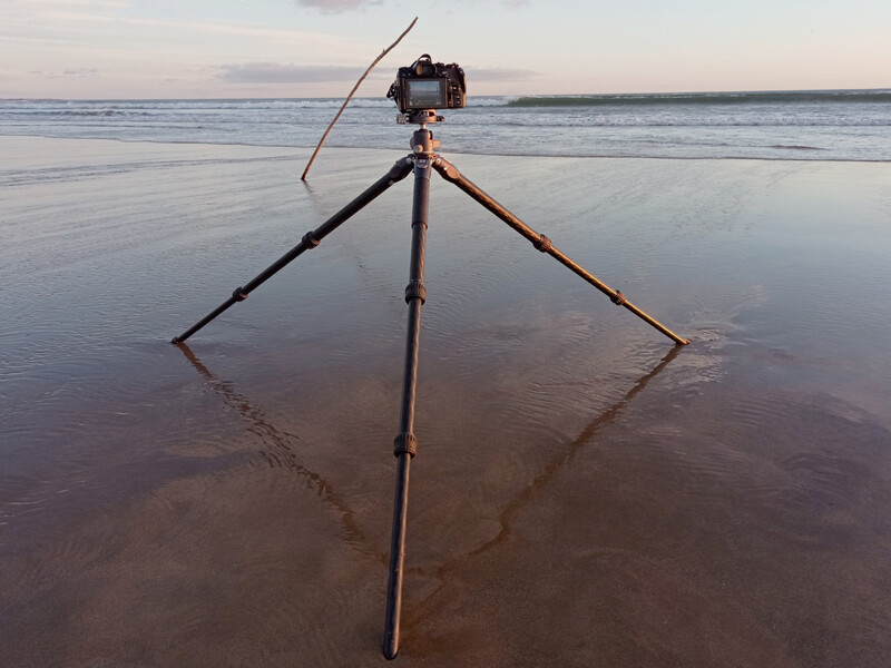 Camera on tripod with extended legs planted in shallow beach water at low tide.