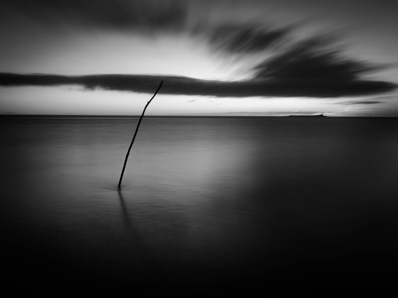 Single curved wooden stake protruding from still, dark water at dusk with dramatic cloudy sky.